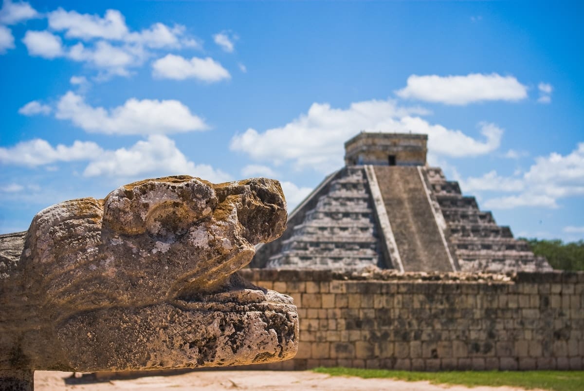 A stone serpent head sculpture in the foreground with the pyramid of Chichen Itza in the background under a blue sky with clouds.